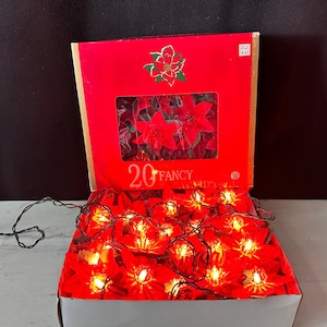 May include: A set of 20 fancy poinsettia-shaped string lights in a red cardboard box. The box features a graphic of a poinsettia flower and the text 20 FANCY. The string lights are illuminated with warm glowing bulbs nestled inside red petals.