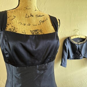 May include: A navy blue satin dress with a square neckline is displayed on a mannequin. A matching bolero jacket with a beaded collar hangs nearby. The dress and jacket are formal wear.