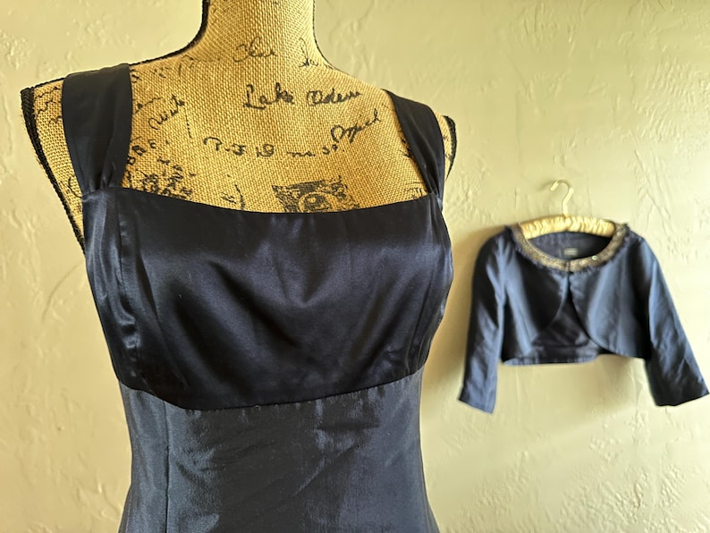 May include: A navy blue satin dress with a square neckline is displayed on a mannequin. A matching bolero jacket with a beaded collar hangs nearby. The dress and jacket are formal wear.