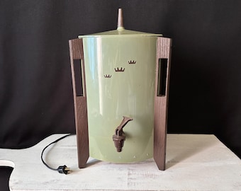 Op de afbeelding: Een saliegroene elektrische drankdispenser met een bruine kunststof kraan en een bruine puntige dekselgreep. De cilindrische behuizing heeft drie kleine kroonvormige uitsparingen en wordt ondersteund door twee verticale bruine houten zijpanelen.