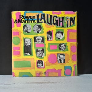 May include: Vintage vinyl record album cover for Rowan & Martin's Laugh-In. The cover is yellow with pink, green, and orange geometric shapes. It features portraits of the cast members and the show's title.