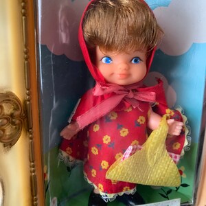 May include: A vintage doll dressed as Red Riding Hood. She is wearing a red hooded dress with a yellow floral pattern and a yellow basket. The doll is displayed in a glass case with a white background.