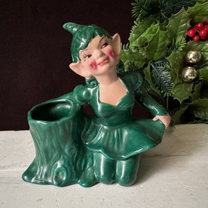 May include: Green ceramic elf figurine with a green dress and pointy ears. The elf is sitting on a green tree stump planter.