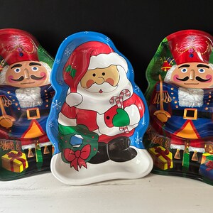 May include: Three Christmas-themed ceramic serving dishes. One features a Santa Claus holding a candy cane and a wreath, another features a Nutcracker holding a sword, and the third features a Nutcracker holding a sword. All three dishes have a green, red, and blue color scheme.