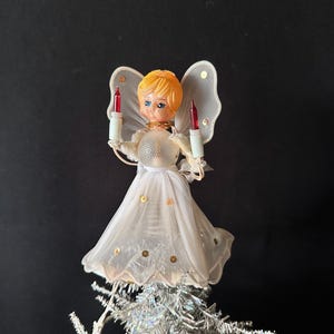 May include: A Christmas tree topper featuring an angel with blonde hair, blue eyes, and a white sheer dress adorned with gold sequins. The angel has translucent wings and holds two white candles with red tips in its hands, positioned above a silver tree.