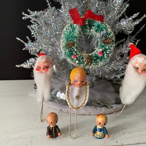 May include: A vintage Christmas tree ornament set featuring a white-bearded Santa, an angel with a halo, and two small figures. The ornaments are made of plastic and have painted details. The set is displayed on a silver tinsel tree with a green wreath.