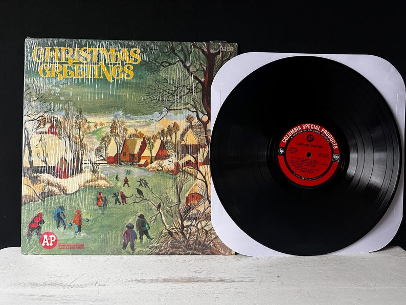 May include: A vintage vinyl record with a winter scene on the cover. The cover art features a snowy village with houses and trees. The record is titled "Christmas Greetings" and is a Columbia Special Products release.