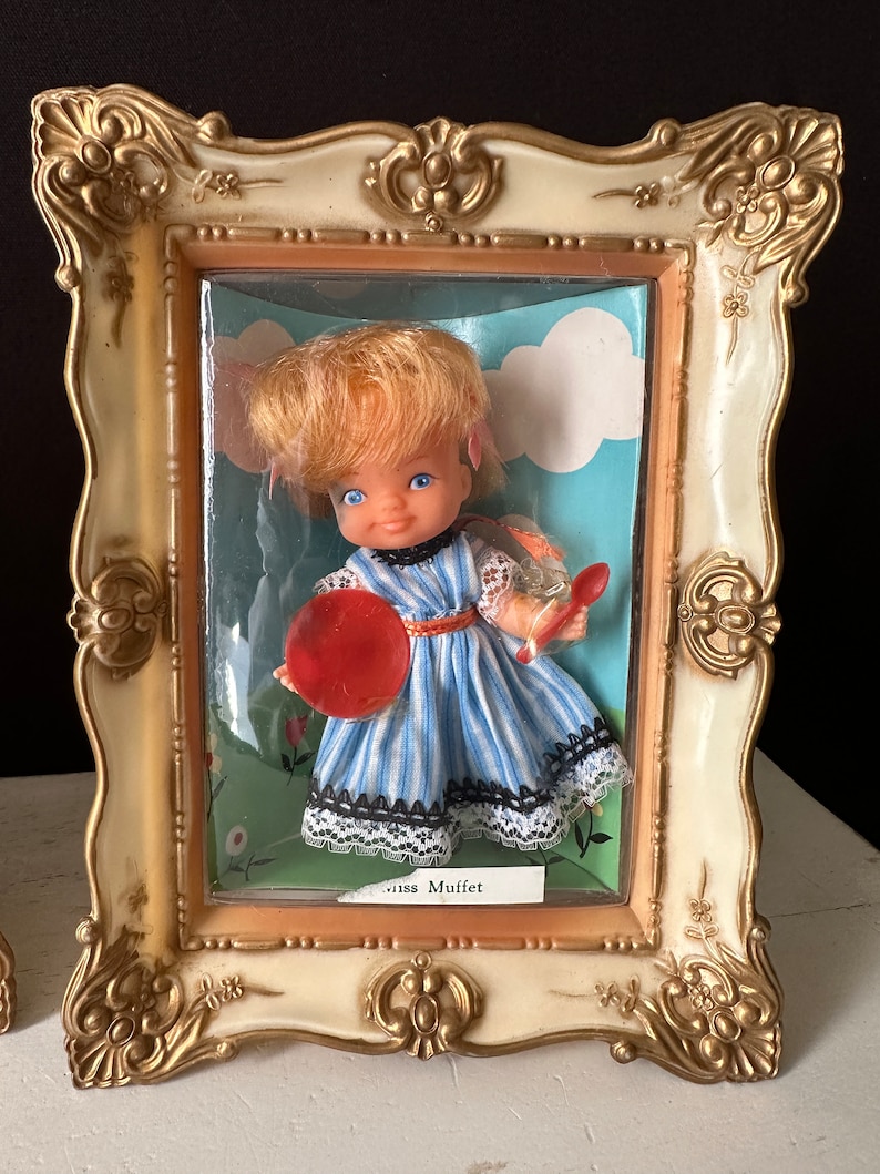 May include: A vintage doll in a blue and white striped dress with a red and white polka dot hat, holding a red plate. The doll is in a plastic frame with a white background and the words "Miss Muffet" printed below.