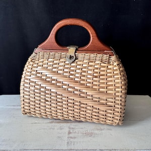 Peut inclure: Un sac à main structuré composé d'un corps en osier tressé marron clair et d'une poignée arquée en bois marron foncé. Le sac est fermé au centre par un fermoir métallique qui relie la poignée en bois à la structure en osier tressé.