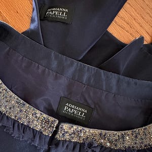 May include: Close-up of a navy blue dress with a beaded neckline and the brand label "Adrianna Papell Occasions". The dress has a satin finish and a ruffled trim along the neckline. The zipper is partially visible.