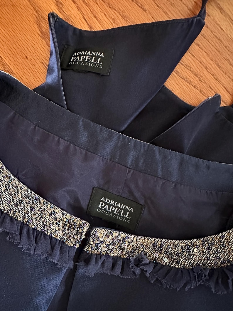 May include: Close-up of a navy blue dress with a beaded neckline and the brand label "Adrianna Papell Occasions". The dress has a satin finish and a ruffled trim along the neckline. The zipper is partially visible.