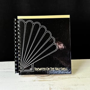 Pode incluir: Um caderno com espiral preto com um desenho de leque branco e o texto "Tidewater on the Half Shell". A borda superior é de cor bege claro. O caderno está sobre uma superfície branca sobre um fundo preto.