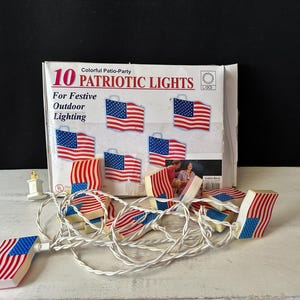Puede incluir: Una cadena de 10 luces patrióticas con forma de banderas americanas, con rayas rojas y blancas y campos de estrellas azules. La caja dice "10 Patriotic Lights For Festive Outdoor Lighting."