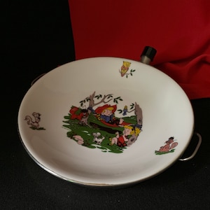 以下が含まれることがあります： 銀色の縁取りと片側に取っ手のある白いエナメル製のボウル。ボウルには、森の風景の中で子供と動物が描かれた漫画のイラストが描かれています。