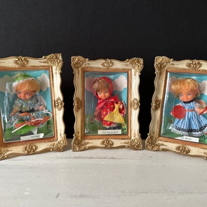 May include: Three vintage doll figurines displayed in ornate gold frames. Each doll is dressed in a different outfit: a green and white dress, a red dress with a yellow basket, and a blue and white striped dress. The frames have a white background and the dolls are displayed against a white cloud background.