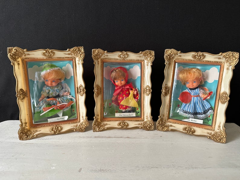 May include: Three vintage doll figurines displayed in ornate gold frames. Each doll is dressed in a different outfit: a green and white dress, a red dress with a yellow basket, and a blue and white striped dress. The frames have a white background and the dolls are displayed against a white cloud background.
