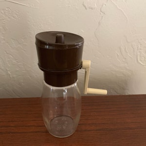 Peut inclure: Un moulin à café manuel brun et beige avec un pot en verre transparent. Le moulin a une manivelle et un couvercle amovible.