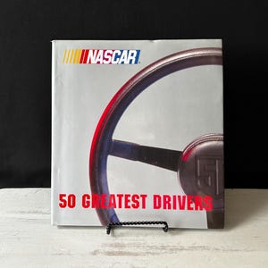 May include: A book cover featuring the NASCAR logo and a partial image of a steering wheel. The title "50 Greatest Drivers" is displayed in red text. The cover is displayed on a black metal stand.