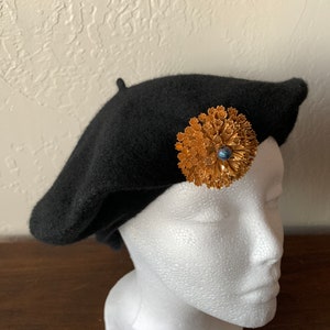 Può includere: Un berretto nero con una spilla a forma di fiore dorata con una pietra blu al centro.