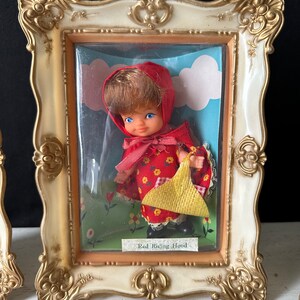 May include: A vintage framed doll figurine of Red Riding Hood wearing a red hooded dress and carrying a yellow basket. The doll is displayed in a plastic case with a blue and white background.