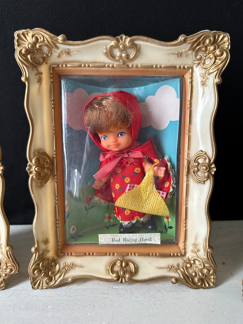 May include: A vintage framed doll figurine of Red Riding Hood wearing a red hooded dress and carrying a yellow basket. The doll is displayed in a plastic case with a blue and white background.