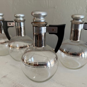May include: Four clear glass decanters with silver accents and black handles. Each decanter has a round body, a silver-colored stopper, and a silver band around the middle. The handles are black and attached to the top.