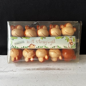 Può includere: Una confezione di plastica trasparente contiene una stringa luminosa con dieci luci a forma di scimmia. Le luci sono marroni e beige, con una faccia di scimmia sorridente. La confezione riporta la scritta "10 ct String Light".