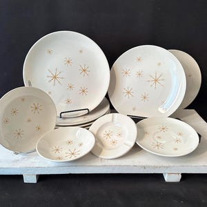 Puede incluir: Un conjunto de vajilla de cerámica blanca con un diseño de estrellas doradas. El juego incluye platos, cuencos y un cenicero. Los platos están dispuestos sobre una tabla de servir de madera blanca. Los platos miden aproximadamente 25 cm de diámetro.