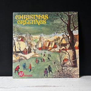 May include: A vintage Christmas record album cover with a snowy winter scene. The cover features a small village with houses, trees, and people walking in the snow. The text "Christmas Greetings" is printed in yellow on a green background.