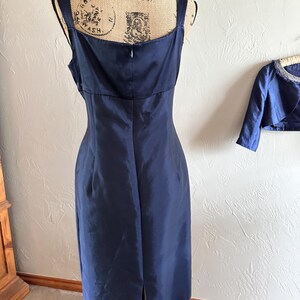 May include: A navy blue, knee-length dress with a square neckline and spaghetti straps. The dress has a fitted bodice and a slight A-line skirt with a slit in the back. A matching bolero jacket is visible in the background.