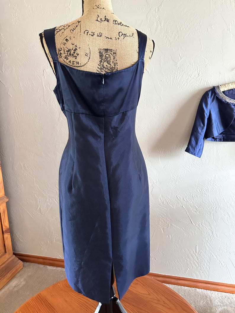May include: A navy blue, knee-length dress with a square neckline and spaghetti straps. The dress has a fitted bodice and a slight A-line skirt with a slit in the back. A matching bolero jacket is visible in the background.