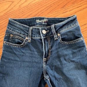 Pode incluir: Close-up de jeans Wrangler em denim azul. Os jeans t&ecirc;m fecho de bot&atilde;o, bolsos frontais e costuras decorativas. A marca "Wrangler" &eacute; vis&iacute;vel na cintura.