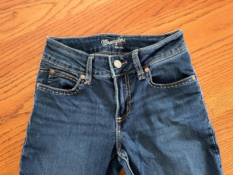 Pode incluir: Close-up de jeans Wrangler em denim azul. Os jeans t&ecirc;m fecho de bot&atilde;o, bolsos frontais e costuras decorativas. A marca "Wrangler" &eacute; vis&iacute;vel na cintura.