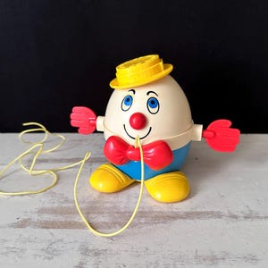 May include: A vintage Humpty Dumpty pull-along toy. The toy is egg-shaped with a yellow hat, red bow tie, and red hands. It has blue eyes, a red nose, and yellow feet. A yellow string is attached.