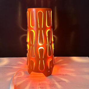 May include: A cylindrical orange table lamp featuring a repeating pattern of vertical, U-shaped cutouts. The lamp emits a warm, glowing light through the openings, casting distinct shadows. It includes a white power cord with an inline switch.