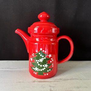 May include: A red ceramic teapot with a lid and handle. The teapot is decorated with a green Christmas tree design, white stars, and white string lights. The teapot is on a white surface with a black background.