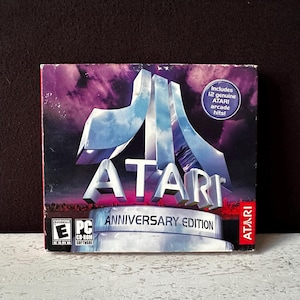 Puede incluir: Una caja de CD para el juego Atari Anniversary Edition. La caja presenta un logotipo de Atari plateado y azul con el texto "ATARI ANNIVERSARY EDITION" en blanco. La caja también incluye el texto "Includes 12 genuine ATARI arcade hits!" en un círculo blanco.