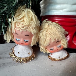 Puede incluir: Dos adornos navideños vintage de ángel con pelo de lana y ojos cerrados. Los ángeles llevan vestidos blancos con ribetes dorados y cuelgan de una bota de Papá Noel roja y blanca.