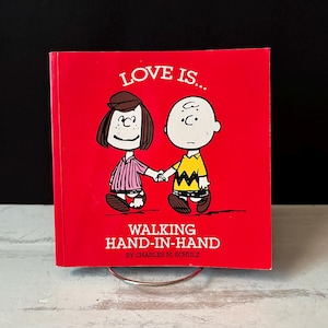 Pode incluir: Um livro vermelho com o texto "LOVE IS... WALKING HAND-IN-HAND" apresenta ilustrações de duas personagens de desenhos animados de mãos dadas. A capa do livro tem um design simples com o nome do autor na parte inferior. O livro é exibido em um suporte de metal prateado.
