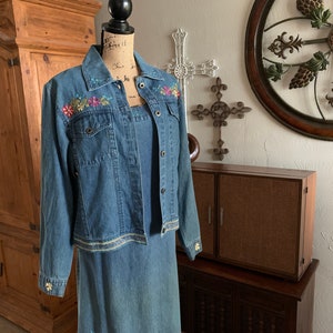 May include: A denim jacket and dress set with floral embroidery. The jacket has a light blue wash and a button-up front. The dress is a matching light blue denim with a straight skirt.