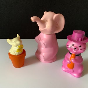 May include: Three pink plastic animal figurines. A pink elephant with a trunk raised in the air, a pink cat wearing a purple hat and holding a carrot, and a white mouse holding a yellow flower in a brown pot.