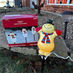 Puede incluir: Una caja de tres marcadores de camino iluminados con forma de muñeco de nieve. La caja es roja y blanca con las palabras "Ashland Christmas Lighted Path Markers" impresas en ella. El muñeco de nieve es amarillo con una bufanda y un gorro rojos. Los marcadores miden 0,9 metros de largo.