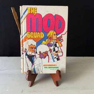 Könnte beinhalten: Ein Vintage-Taschenbuch mit dem Titel "The Mod Squad: Assignment: The Arranger" mit einem bunten Cover, das drei Personen und ein psychedelisches Design zeigt.