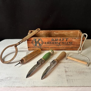 May include: A collection of antique kitchen utensils, including cheese knives, a cheese slicer, and a wooden Kraft Cream Cheese box. The tools have wooden and metal handles, showing signs of age. The box features the Kraft Cream Cheese logo.