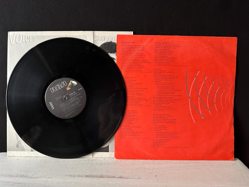 May include: A black vinyl record with a red and white label that says "RCA Victor" and "Voices" by John Oates. The record is on a white background with a red and white album cover that says "Voices" by John Oates.