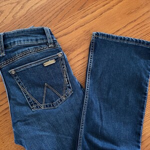 Pode incluir: Jeans azul escuro com um design cl&aacute;ssico de cinco bolsos. Apresenta uma etiqueta met&aacute;lica da marca no bolso traseiro e costuras decorativas. Os jeans est&atilde;o colocados sobre uma superf&iacute;cie de madeira.
