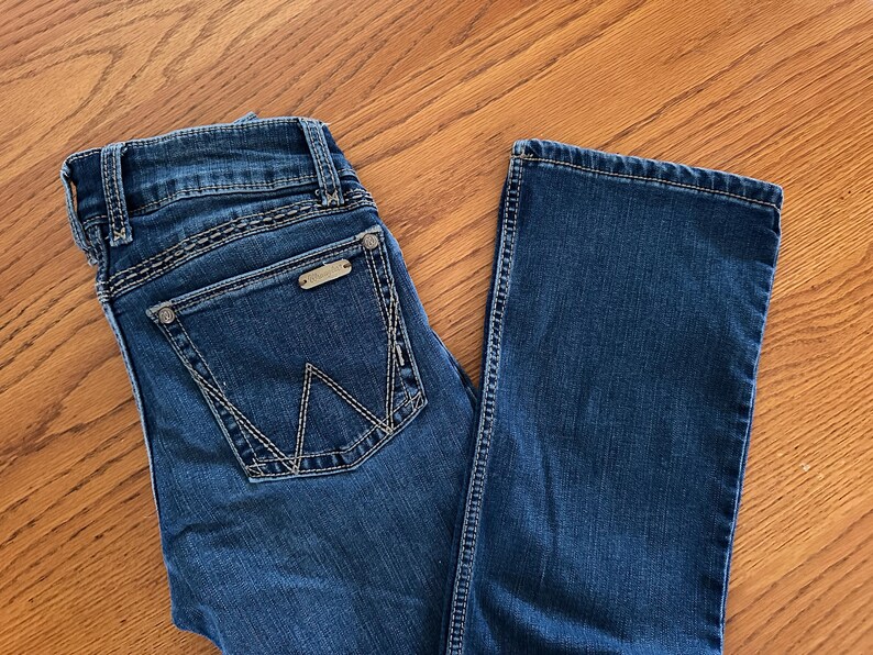 Pode incluir: Jeans azul escuro com um design cl&aacute;ssico de cinco bolsos. Apresenta uma etiqueta met&aacute;lica da marca no bolso traseiro e costuras decorativas. Os jeans est&atilde;o colocados sobre uma superf&iacute;cie de madeira.