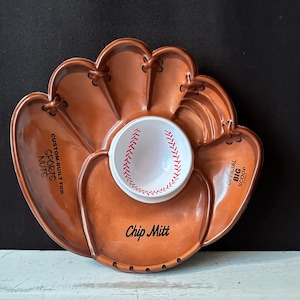 May include: A baseball-themed chip and dip serving dish. The brown ceramic dish is shaped like a baseball glove with a white bowl in the center, decorated with red stitching. Text on the dish includes "Custom Built for Sports Nuts", "Official Big Scoop", and "Chip Mitt".