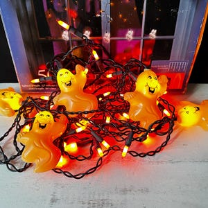 May include: A string of orange Halloween ghost lights with black wire. The ghosts are smiling and have a glow in their eyes. The lights are strung together with a black wire and have orange bulbs.