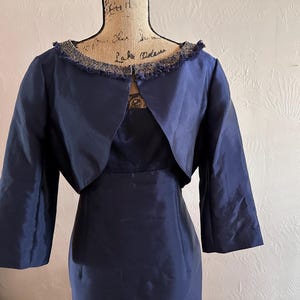 May include: A navy blue dress with a matching bolero jacket. The dress has a fitted silhouette, and the jacket features three-quarter sleeves and a decorative trim around the neckline. The outfit is displayed on a mannequin.